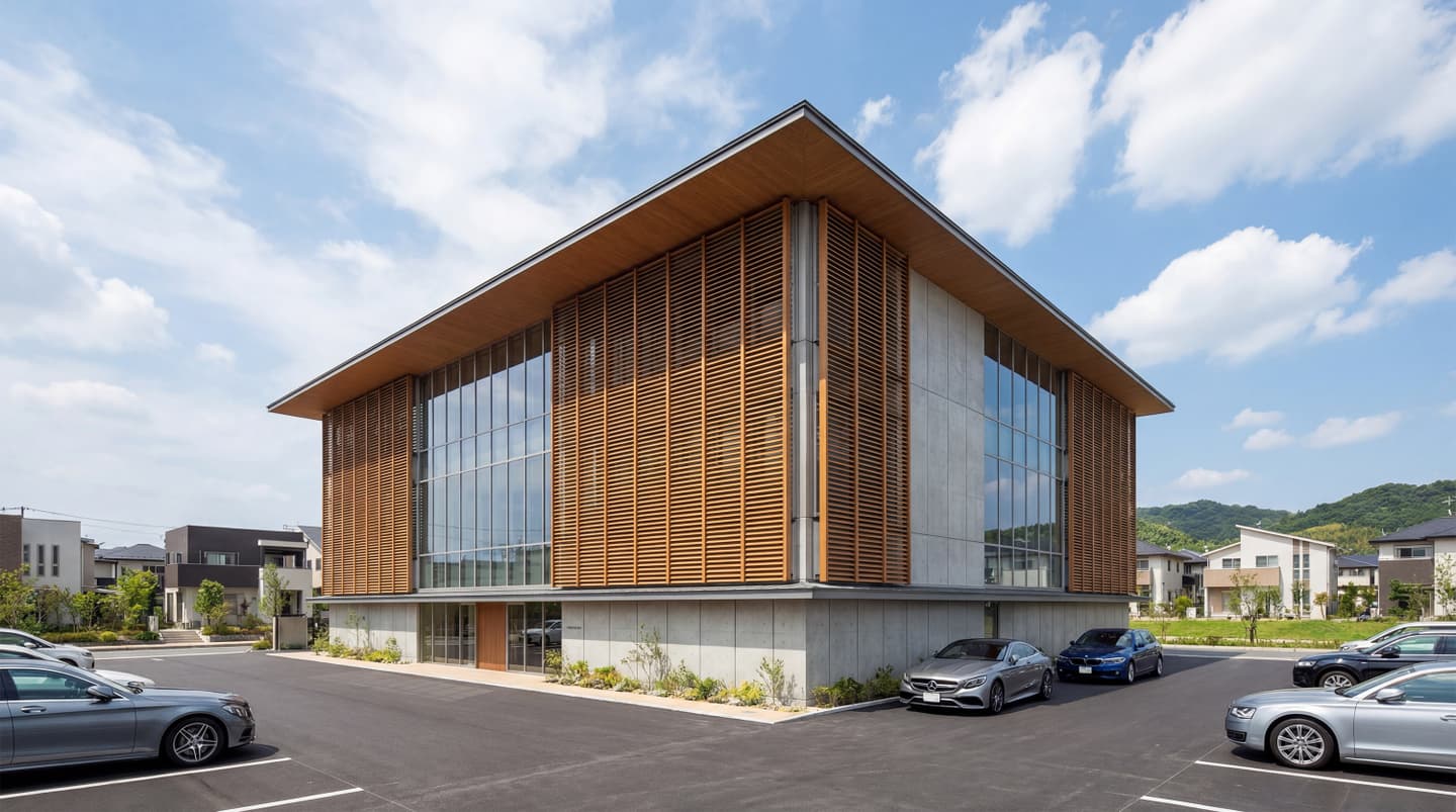 青空の下に建つ、木製ルーバーとコンクリート打ち放し壁面が特徴的な2階建てモダンオフィスビルの外観パース。大きなガラス窓と平らな庇（ひさし）を持つ現代的なデザイン。建物の前には舗装された駐車場があり、数台の乗用車が止まっている。生成AIによる制作画像。