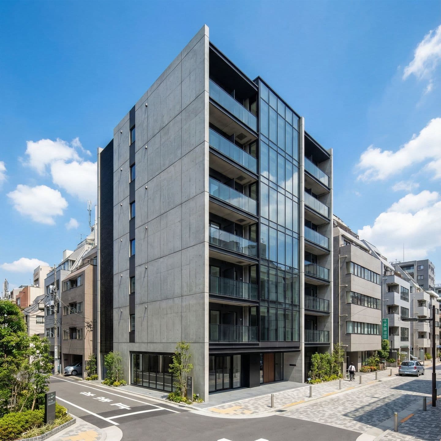 晴天の青空の下、街角に建つモダンな8階建てマンションの外観。コンクリート打ち放しの壁面と、空を反射する大きなガラス張りの窓、黒い金属フレームが特徴的な現代建築デザイン。