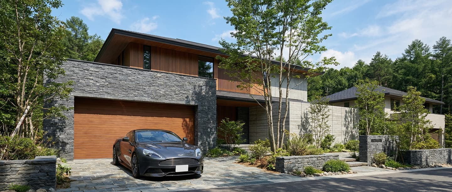 3DCADと生成AIで制作した、石張りの外壁と木製ガレージドアが特徴的なモダンな高級住宅の外観パース。手前の石畳のドライブウェイにはグレーのスポーツカーが駐車されており、豊かな緑に囲まれている。